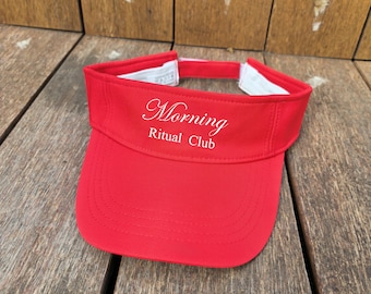 May include: A bright red sun visor featuring the white embroidered text Morning Ritual Club on the front panel. The visor has a curved brim with visible stitching and an adjustable strap at the back with a white interior lining.