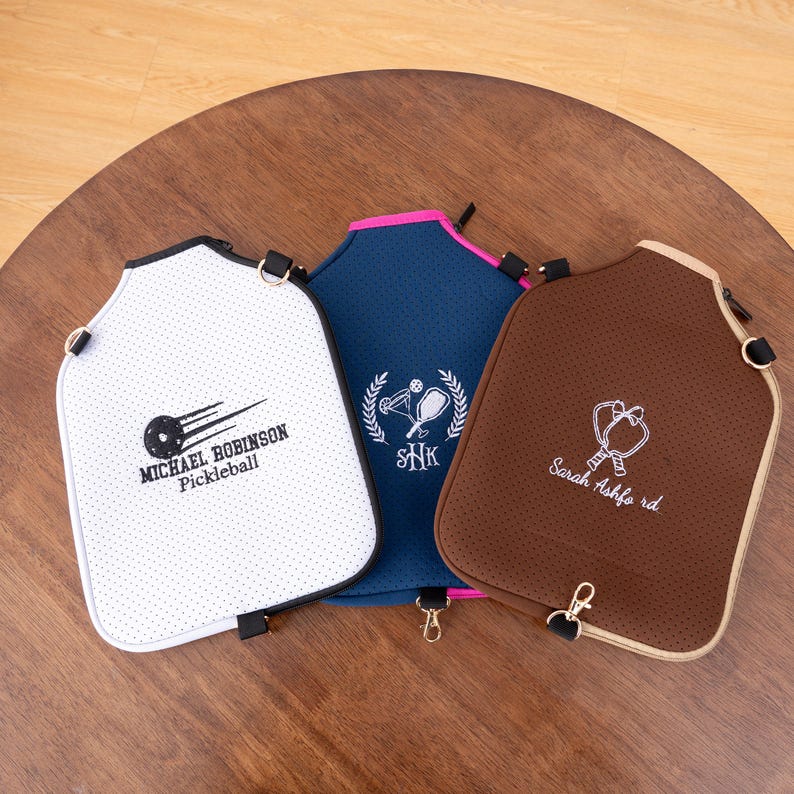 May include: Three pickleball paddle covers in white, navy blue, and brown. Each cover has a unique design and text, including the names "Michael Robinson Pickleball" and "Sarah Ashford rd."
