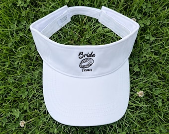 May include: A white sun visor featuring the embroidered black text "Bride Team" above a small black line drawing of an oyster shell. The visor has a structured front panel, a curved brim with stitching, and an adjustable strap at the back.