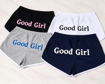 Puede incluir: Cuatro pares de pantalones cortos de estilo deportivo con ribete blanco, cada uno con el texto "Good Girl" impreso en la parte posterior. Los pantalones vienen en negro con texto rosa, blanco con texto negro, gris con texto azul y azul marino con texto blanco.
