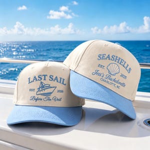 Peut inclure: Deux casquettes de baseball crème avec des visières bleu clair. La casquette de gauche présente un motif de bateau brodé en bleu avec le texte LAST SAIL EST. 2026 Before The Veil. Celle de droite porte un coquillage avec SEASHELLS EST. 2025 Jess's Bachelorette CHARLOTTE, NC.