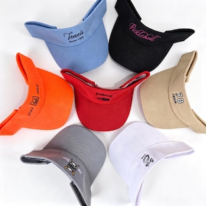 Può includere: Una collezione di sette visiere parasole in vari colori tra cui azzurro, nero, arancione, rosso, beige, grigio e bianco. Ogni visiera presenta testi o grafiche ricamate uniche come Tennis Social Club, Pickleball, Stay Wild, 78 Jason e Bride.