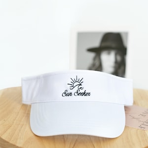 May include: White visor with a curved brim. The visor features the embroidered text "Sun Seeker" in black, along with a sun and wave graphic. The visor is designed to shield the eyes from the sun.