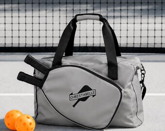 Puede incluir: Una bolsa de pickleball gris claro con asas y bordes negros, que presenta un bolsillo frontal con un gráfico de pala de pickleball negra y el texto CHRISTABELLE. Dos palas negras están insertadas en el lateral y dos pelotas naranjas cerca.