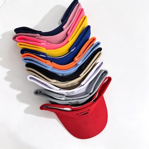 Könnte beinhalten: Ein Stapel von elf Sonnenblenden in verschiedenen Farben, darunter Rot, Grau, Weiß, Beige, Schwarz, Hellblau, Orange, Dunkelblau, Gelb, Pink und Marineblau. Jede Sonnenblende hat einen geschwungenen Schirm und einen verstellbaren Riemen mit Stickerei.