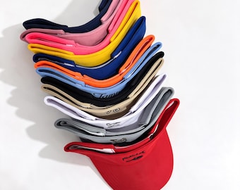May include: A stack of eleven sun visors in various colors including red, grey, white, beige, black, light blue, orange, dark blue, yellow, pink, and navy. Each visor features a curved brim and an adjustable strap, with some displaying embroidered text.