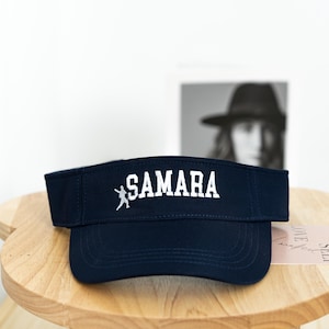May include: Navy blue visor with the name "SAMARA" embroidered in white, along with a small white figure. The visor is sitting on a wooden table. A black and white photo is in the background.