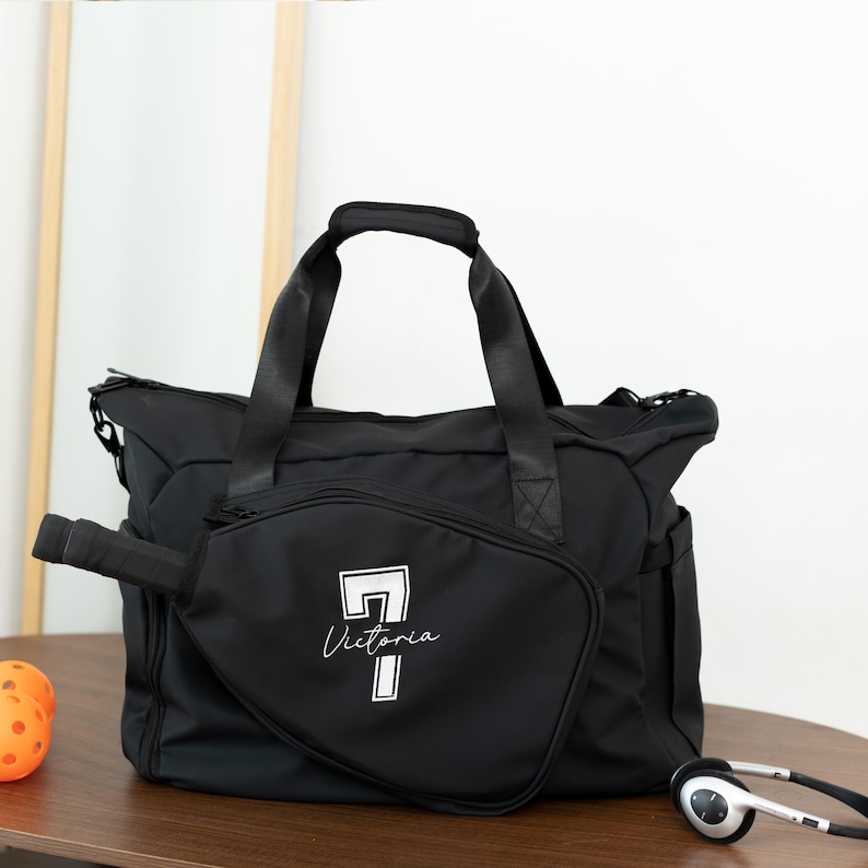 May include: Black duffel bag with a white graphic of a pickleball paddle and the name "Victoria". The bag has a top handle and shoulder straps. A pickleball paddle and headphones are next to the bag.
