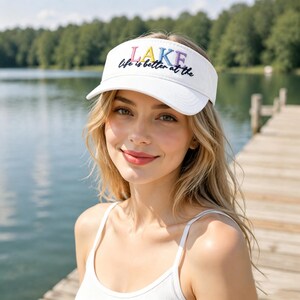 May include: A white visor featuring the embroidered text "life is better at the" in black script and the word "LAKE" in large, multicoloured block letters. The visor has a curved brim and an adjustable strap, providing a clean, simple design.