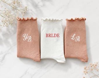 May include: Three pairs of socks with ruffled cuffs. Two socks are dusty rose with white cursive initials YB and LR printed on them. The center sock is white with the red text BRLDE printed in a serif font. The socks are arranged side by side.