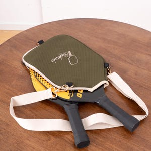 May include: Olive green pickleball paddle cover with a white strap and two pickleball paddles. The cover has the name "Stefanie" and a pickleball graphic. The paddles are black with yellow and white accents.