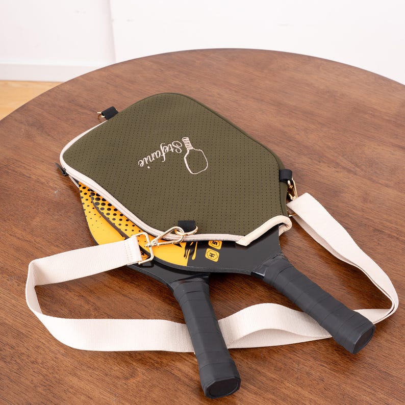 May include: Olive green pickleball paddle cover with a white strap and two pickleball paddles. The cover has the name "Stefanie" and a pickleball graphic. The paddles are black with yellow and white accents.