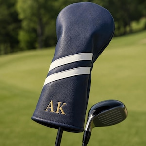 May include: Navy blue golf club head cover with two white stripes and gold embroidered initials "AK". The cover is on a golf club with a black shaft and a shiny black club head. The background is a green golf course.
