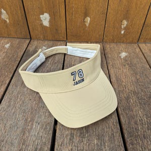 Op de afbeelding: Een beige zonneklep met het getal 78 geborduurd in marineblauw en wit, met de naam JASON in marineblauw borduurwerk eronder. De zonneklep heeft een witte binnenband en een verstelbare sluiting aan de achterzijde voor een goede pasvorm.