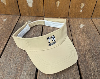 May include: A beige visor featuring the number 78 embroidered in navy blue and white, with the name JASON embroidered in navy blue underneath. The visor has a white interior headband and an adjustable strap closure at the back for a secure fit.