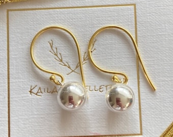 May include: A pair of silver ball earrings with gold hooks. The earrings are on a white card with the text "Kaila" and "Ellet" printed on it.