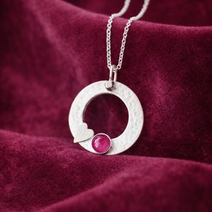 May include: A silver pendant necklace with a textured, circular design. A small heart shape is on the left side of the circle, and a round, pink gemstone is set at the bottom. The necklace hangs on a silver chain against a deep red velvet background.
