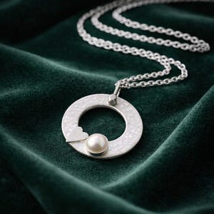 May include: A silver pendant necklace with a hammered texture. The pendant is a circle with a small heart and a pearl in the centre. The necklace chain is also silver, displayed on a dark green velvet surface.