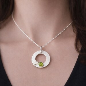 May include: A silver pendant necklace featuring a textured circular design. The pendant includes a small heart and a round, light green gemstone. The necklace is suspended from a delicate silver chain.