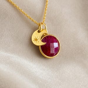 May include: A gold chain necklace featuring a round, faceted deep pink gemstone pendant and a small gold crescent moon charm with an engraved starburst detail. The pendant and charm are suspended from a delicate gold link chain.