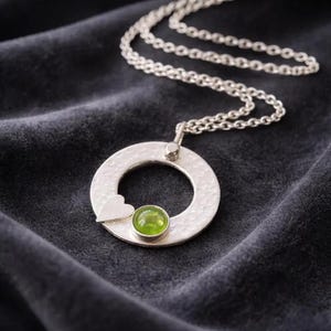 May include: A silver pendant necklace with a hammered texture. The pendant has a circle with a small heart and a round, green gemstone. The silver chain rests on a dark grey fabric.