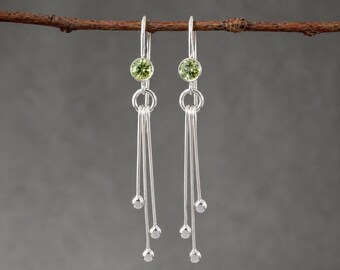 May include: A pair of silver dangle earrings. Each earring features a small, round, light green gemstone at the top, connected to a series of silver wires ending in small silver beads. The earrings are suspended from a thin, brown branch against a gray background.