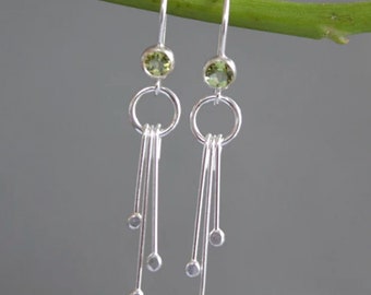 May include: Silver dangle earrings with a green gemstone and a circle design. The earrings have multiple strands of silver with small beads at the ends.