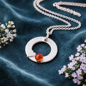 May include: A silver pendant necklace with a hammered texture, featuring a small heart and a round, orange-red gemstone. The necklace hangs on a delicate silver chain, set against a dark teal background with white flowers.