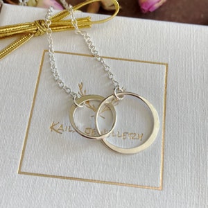 Könnte beinhalten: Eine silberne Halskette mit zwei ineinander verschlungenen Kreisen. Die Halskette liegt auf einem weißen Hintergrund mit dem sichtbaren Text "Kail Jewellery".