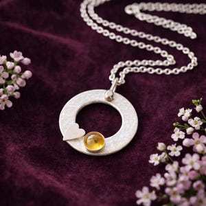 May include: A silver pendant necklace with a hammered texture, featuring a small heart and a yellow gemstone. The necklace hangs on a double chain against a dark purple background, with pink flowers in the corners.