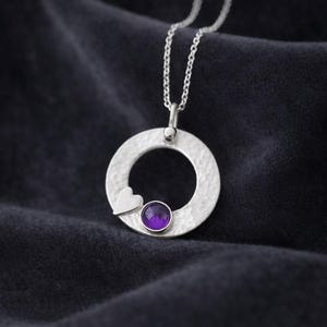 May include: A silver pendant necklace with a hammered circular design, a small heart, and a round, purple gemstone. The necklace is suspended from a fine silver chain. The background is dark.