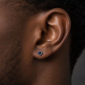 May include: A small, round stud earring with a dark blue and silver design. The earring is set against the skin near the ear. The earring has a concentric circle pattern.