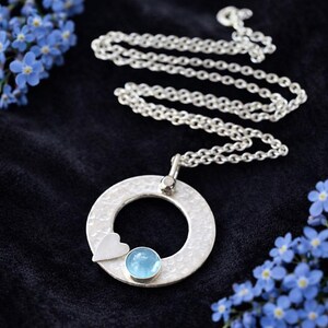 May include: A silver necklace with a textured, circular pendant. The pendant features a small heart and a light blue gemstone. The chain is delicate, and the necklace is displayed on a dark background with blue flowers.
