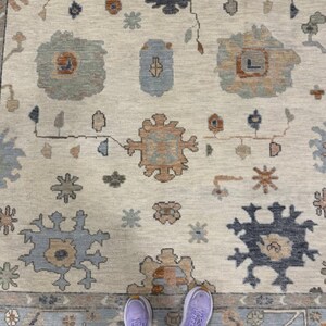 May include: Cream-colored area rug with a detailed floral pattern in shades of blue, tan, and rust. The rug features a border with geometric designs. The image includes a person's feet wearing purple sneakers.