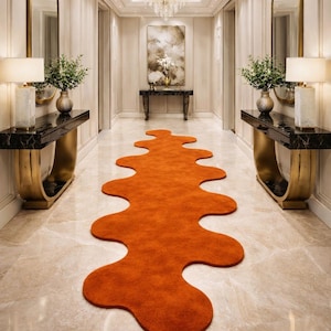May include: An orange, organic-shaped rug runs down the centre of a hallway. The hallway features marble flooring, gold accents, and a chandelier. Two console tables with lamps and mirrors flank the hallway.