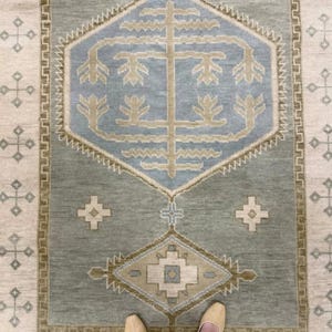 Op de afbeelding: Een rechthoekig vloerkleed met een centraal geometrisch ontwerp in tinten blauw, beige en groen. Het vloerkleed heeft een herhalende patroonrand en een centraal ontwerp met een boomachtig motief. Van bovenaf bekeken.