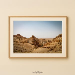 May include: A framed landscape photograph depicting a desert canyon under a clear blue sky. The image shows tan and brown rock formations, with a small amount of green vegetation in the canyon's depths. The photograph is in a light-coloured wooden frame.
