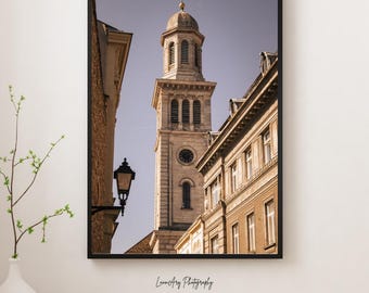 Pode incluir: Fotografia emoldurada de uma alta e ornamentada torre de igreja com relógio, entre edifícios. A imagem está numa moldura preta e apresenta um candeeiro de rua. A fotografia é assinada "LunaArt Photography".