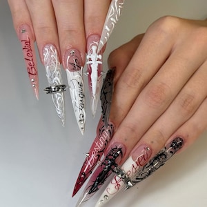 May include: A set of long, pointed acrylic nails with intricate designs. The nails feature a variety of colors, including red, white, and black, with detailed artwork and text such as "Celestial", "Forgive", and "Destiny". Some nails have silver studded rings.