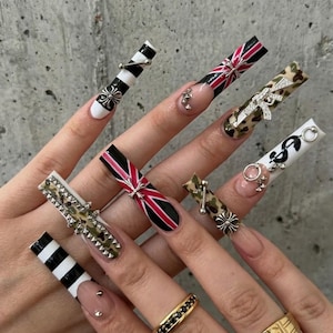 May include: A hand showing long acrylic nails with various designs. These include black and white stripes, camouflage, a Union Jack, and dollar signs. Silver charms and studs are used as embellishments. Some nails have a clear base.