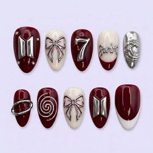 May include: A collection of ten artificial nails in a deep red and white colour scheme. The nails feature silver embellishments, including bows, the number 7, and a wave design. Some nails have the word "swim" in silver.