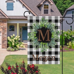 May include: A garden flag with a black and white check pattern, featuring a green wreath design around a brown letter "M". The name "Megan" is printed in brown at the bottom. The flag is displayed in a garden.