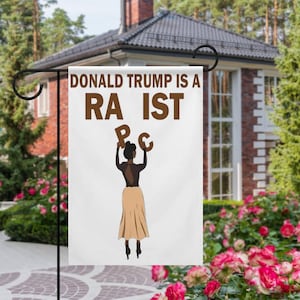 May include: A white garden flag with the text "DONALD TRUMP IS A RA IST" in brown letters. A graphic of a person holding up the letters "P" and "C" is below the text. The flag is hanging in a garden.