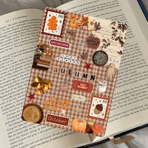May include: A rectangular bookmark with a brown and white plaid pattern featuring various autumn-themed stickers including an orange leaf, a pumpkin, a clock, a ghost, a cup of coffee, and the text "COSY SEASON", "autumn", and "IN MY COZY GIRL ERA".