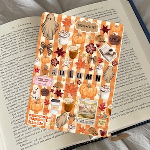 Op de afbeelding: Een decoratieve stickerpagina met herfstthema met pompoenen, koffiekopjes, geesten en herfstbladeren. Het woord "AUTUMN" is in zwarte letters geschreven. De pagina ligt op een open boek.