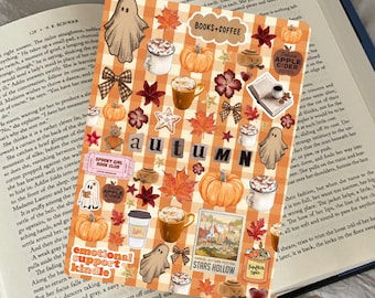 May include: A decorative autumn-themed sticker sheet with pumpkins, coffee cups, ghosts, and fall leaves. The word "AUTUMN" is spelled out in black letters. The sheet is placed on top of an open book.