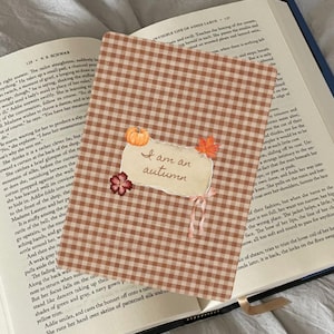 May include: A rectangular bookmark with a brown and white gingham pattern featuring a central torn-paper label with the handwritten text "I am an autumn." The label is decorated with a small orange pumpkin, a red maple leaf, and a flower.