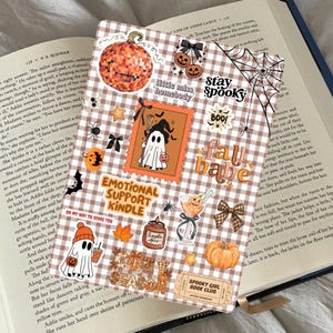 Op de afbeelding: Een rechthoekige Kindle-skin met een bruin-wit ruitpatroon, versierd met diverse herfststickers zoals een discopompoen, een geest met een heksenhoed, een spinnenweb en de tekst Emotional Support Kindle.