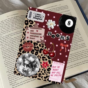 Puede incluir: Una cubierta de libro decorativa con un fondo de estampado de leopardo, adornada con pegatinas, una bola de discoteca, cerezas y la palabra "BOOKS". Incluye el texto "one more chapter" y "Buy me books and call me pretty."