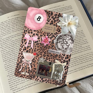 Könnte beinhalten: Eine Collage auf einer Buchseite mit einer rosa Achtkugel, einem Cocktail, einer Discokugel, einer Kamera und den Worten "I love you". Der Hintergrund ist ein Leopardenmuster mit einer rosa Schleife und einem Ticket.